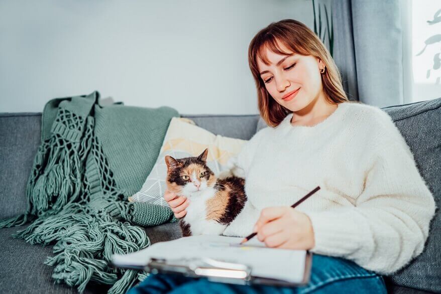 Frau auf Sofa mit Katze praktiziert Selbstreflexion und schreibt in Notizbuch