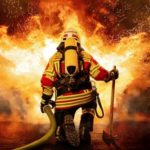 Ein Feuerwehrmann hält einen Löschschlauch und kniet vor einem großen Feuer.
