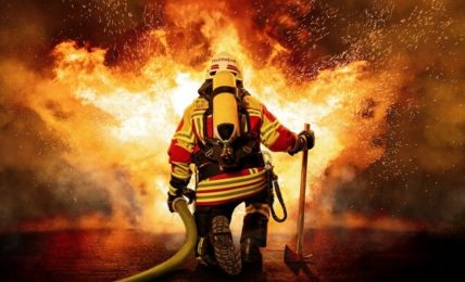 Ein Feuerwehrmann hält einen Löschschlauch und kniet vor einem großen Feuer.
