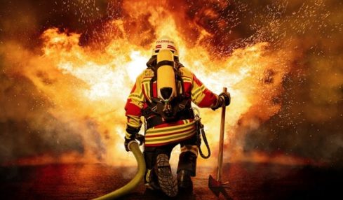 Ein Feuerwehrmann hält einen Löschschlauch und kniet vor einem großen Feuer.