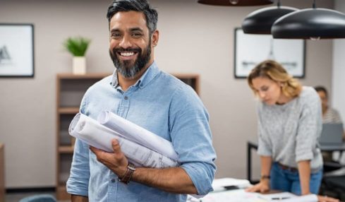 Ein lächelnder, schwarzhaariger angehender Bauzeichner mit Bauplänen im Arm während der Ausbilung