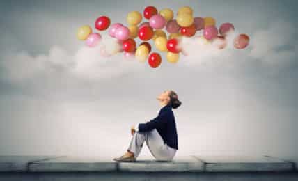 Frau sitzt auf Mauer und guckt zu bunten Luftballons über sich im Himmel