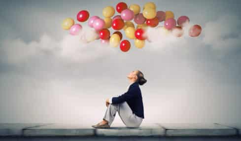 Frau sitzt auf Mauer und guckt zu bunten Luftballons über sich im Himmel