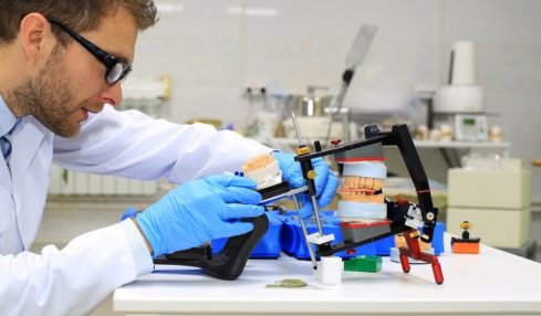 Ein Zahntechniker mit schwarzer Brille, weißem Kittel & blauen Hygienehandschuhen beim Überprüfen seiner Arbeit