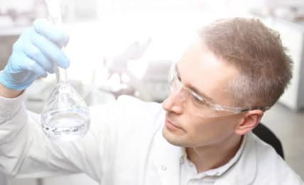 Ein Stoffprüfer mit Schutzbrille, weißem Laborkittel und Reagenzglas in der Hand.