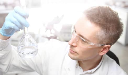 Ein Stoffprüfer mit Schutzbrille, weißem Laborkittel und Reagenzglas in der Hand.