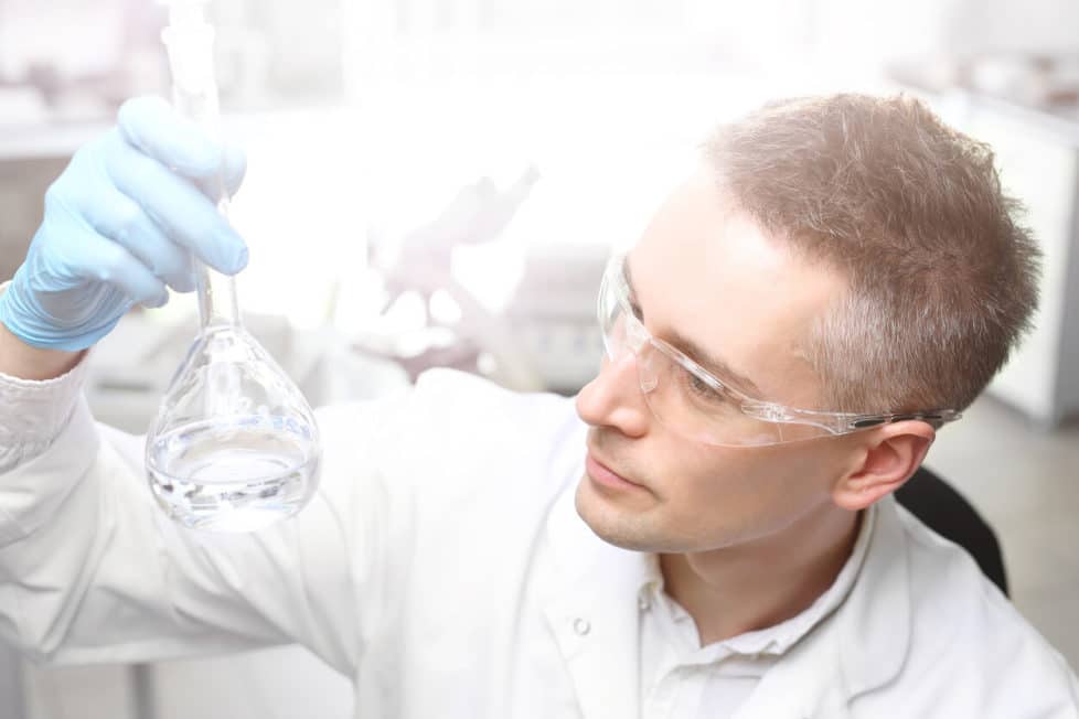 Ein Stoffprüfer mit Schutzbrille, weißem Laborkittel und Reagenzglas in der Hand.