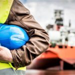 Ein blauer Helm im Arm eines Schiffmechanikers. Im Hintergrund ist ein großes Frachtschiff.