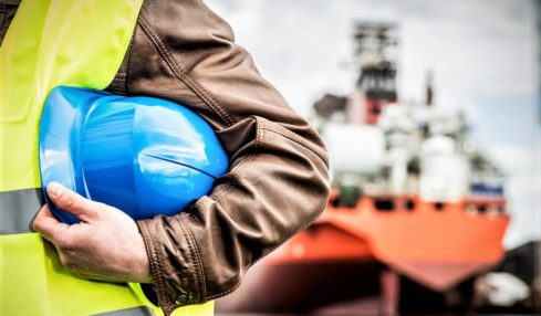 Ein blauer Helm im Arm eines Schiffmechanikers. Im Hintergrund ist ein großes Frachtschiff.