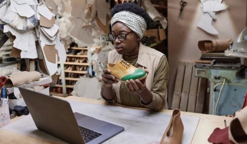 Ein dunkelhäutige Schuhmacherin präsentiert einen Schuh am Laptop.