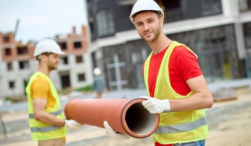 2 angehende Tiefbaufacharbeiter in der Ausbildung halten ein großes Plastikrohr auf der Baustelle