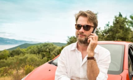 Mann telefoniert an Auto gelehnt im Urlaub mit Smartphone