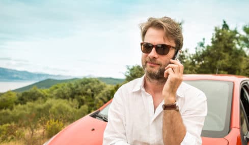Mann telefoniert an Auto gelehnt im Urlaub mit Smartphone