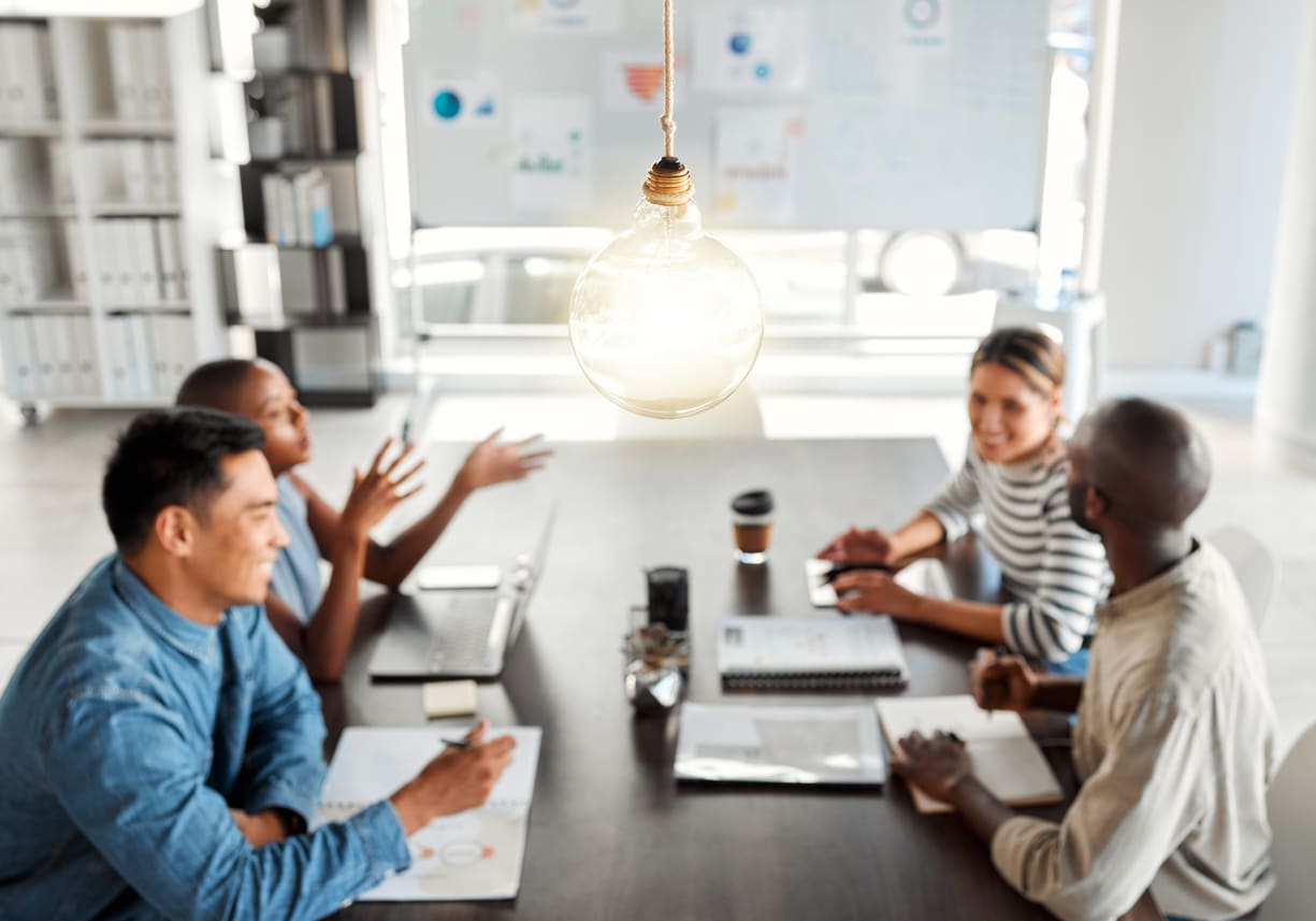 Vier Kollegen sitzen in hellem Büro am Schreibtisch und diskutieren unter großer Lampe