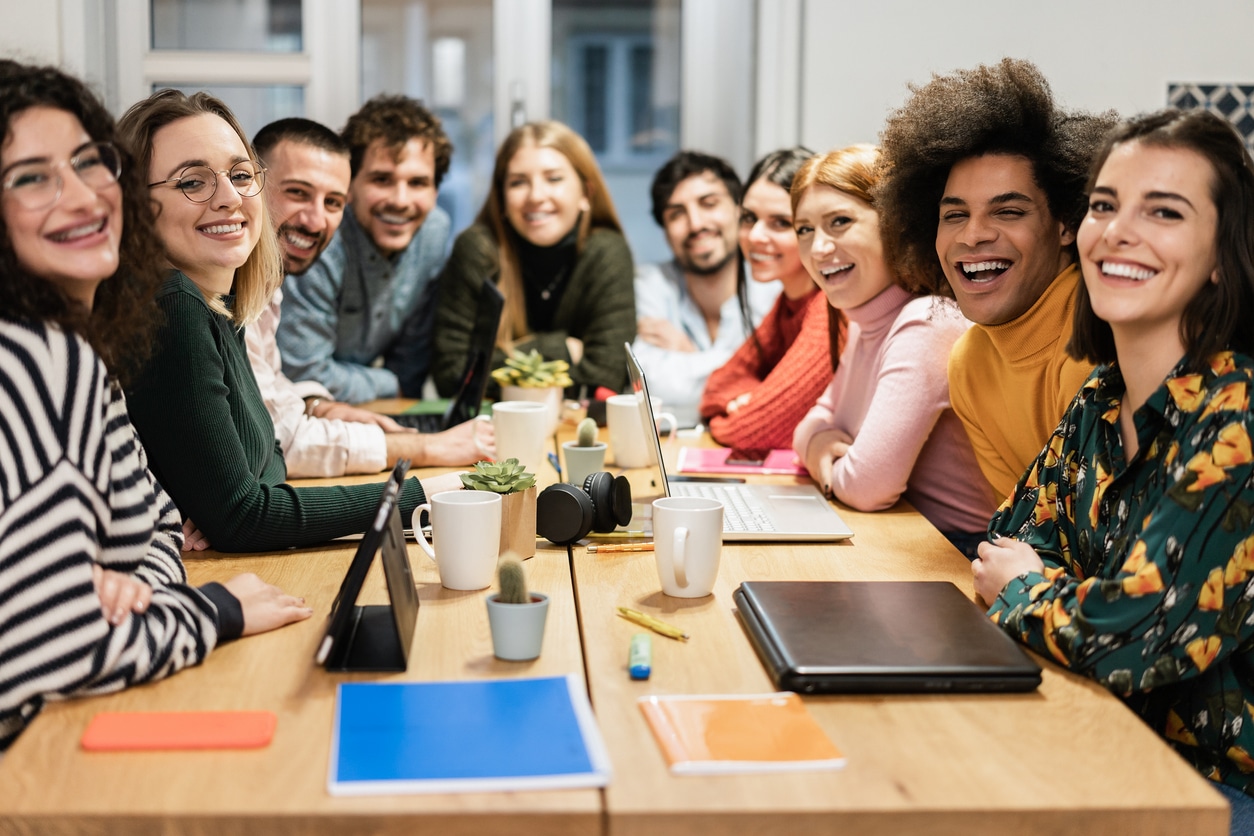 Junge Mitarbeiter sitzen im Büro am Konferenztisch vor Laptops und lächeln in die Kamera