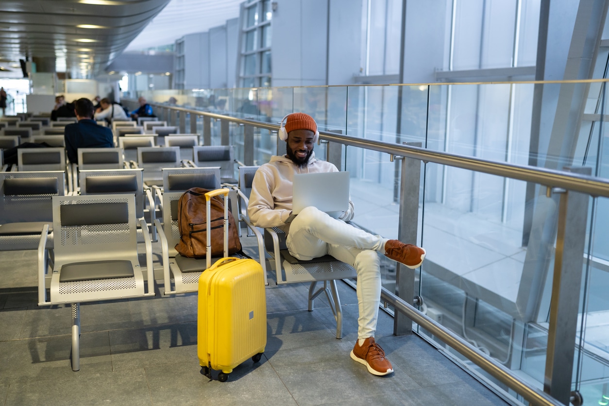 Junger Mann der Generation Z sitzt am Flughafen mit Koffer und aufgeschlagenem Laptop auf dem Schoß