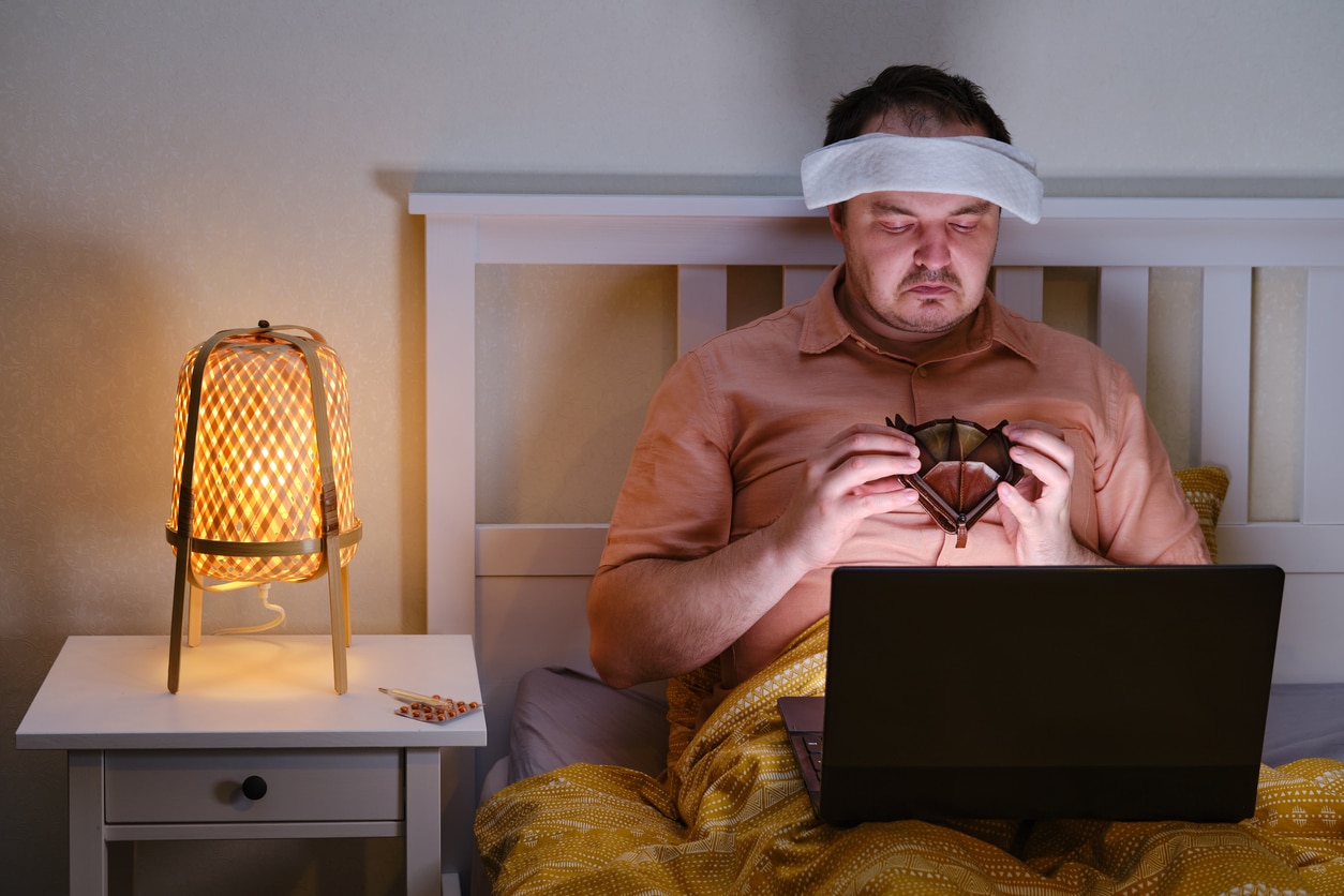 Lohnfortzahlung: Kranker Mann im Bett mit Laptop blickt traurig in seine leere Geldbörse