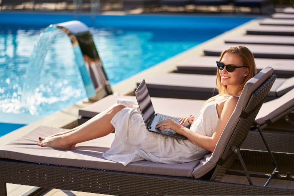 Junge Frau liegt mit Laptop auf dem Schoß am Pool - Homeoffice im Ausland