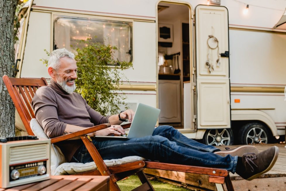 Grauhaariger attraktiver Mann liegt vor seinem Wohnmobil mit Laptop auf dem Schoß im Liegestuhl - Homeoffice im Ausland