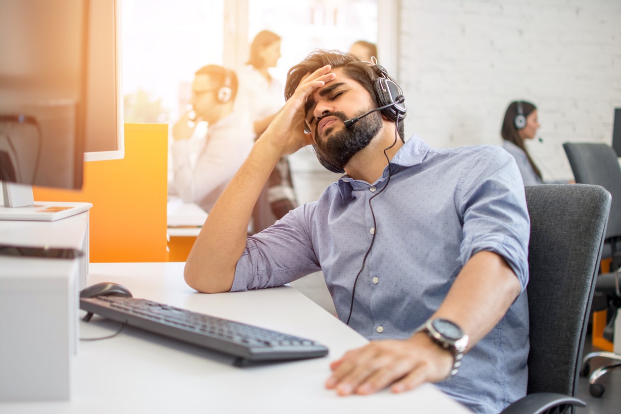 Erschöpfter junger Mann mit Headset am Arbeitsplatz im Großraumbüro