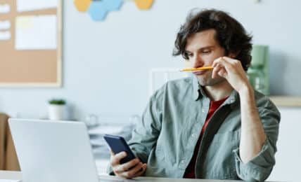 Prokrastination: junger Mann vor Laptop schaut abgelenkt auf Smartphone