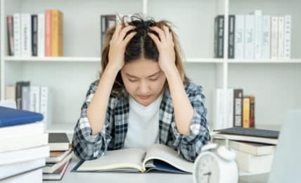 Regelstudienzeit - junge Studentin lernt verzweifelt über Büchern mit Wecker auf dem Tisch