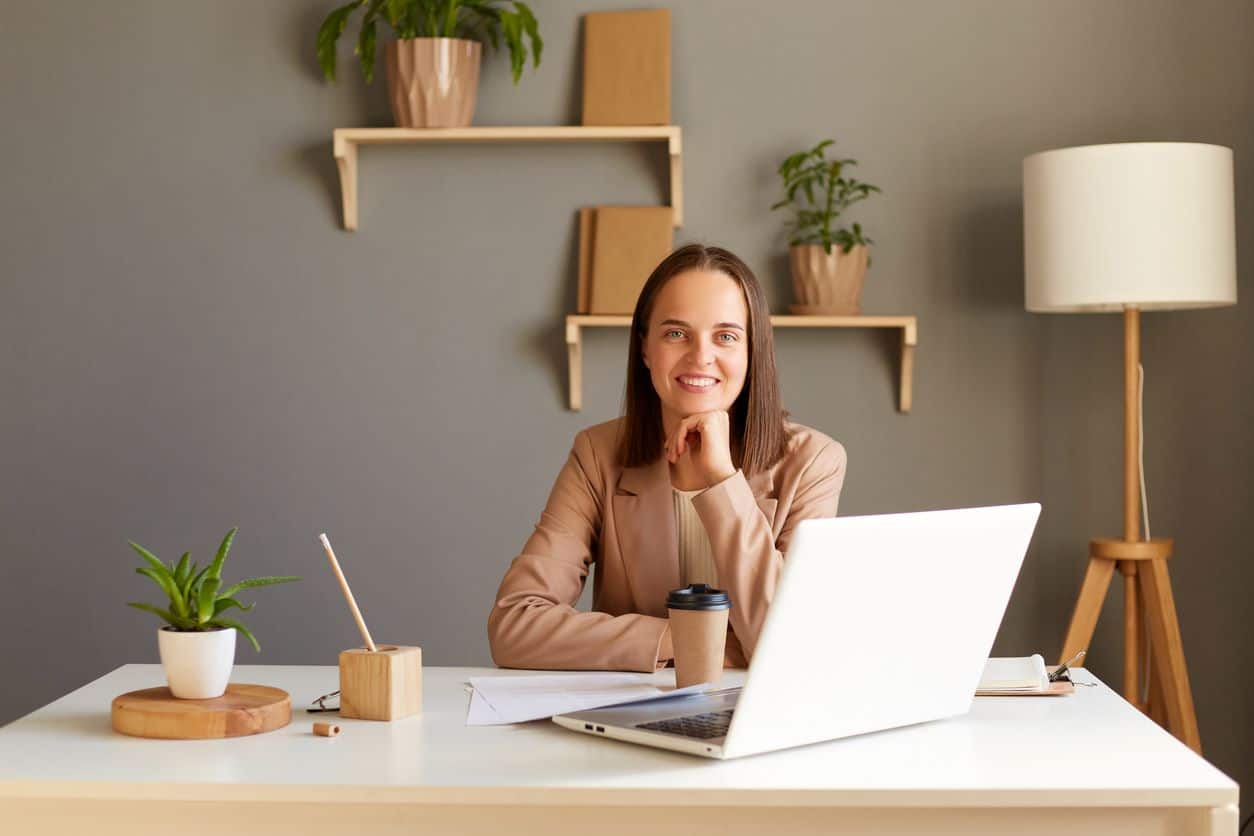 Junge lächelnde, positiv denkende Frau am Schreibtisch im Homeoffice vor Laptop