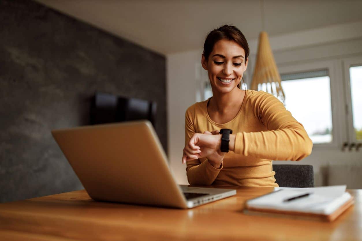 Frau am Laptop blickt lächelnd auf Armbanduhr - kein Zeitdruck am Arbeitsplatz