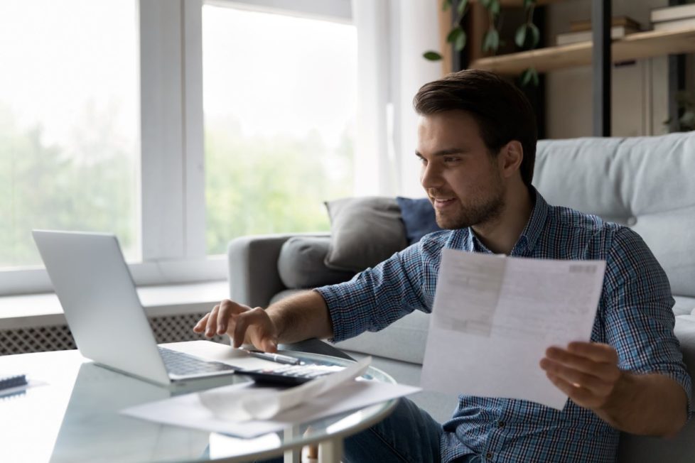 Mann prüft Unterlagen und Gehaltsnachweis zuhause am Laptop und in Papierform