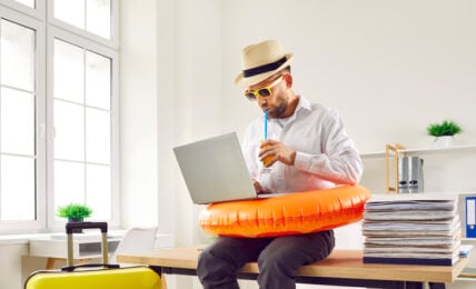 Business man in beach ring and summer hat sits on table in office with holiday suitcase, uses laptop, books online tickets to his annual leave vacation destination, and sips orange juice through straw