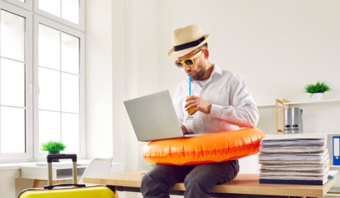 Business man in beach ring and summer hat sits on table in office with holiday suitcase, uses laptop, books online tickets to his annual leave vacation destination, and sips orange juice through straw