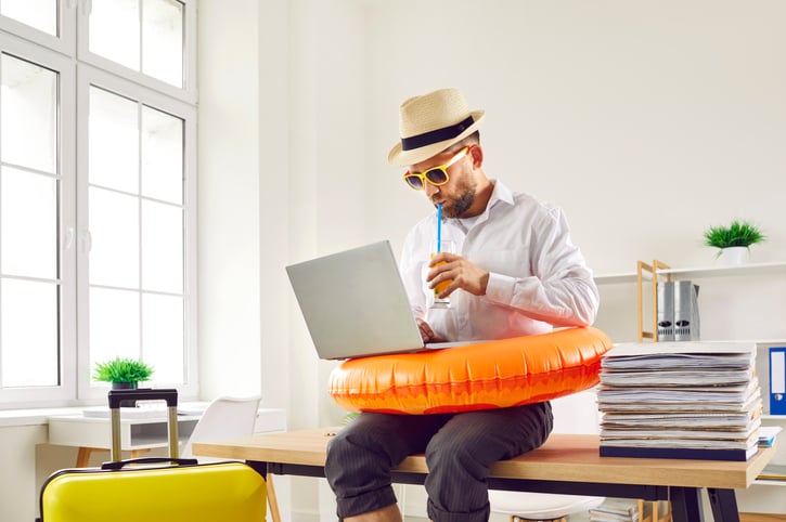 Business man in beach ring and summer hat sits on table in office with holiday suitcase, uses laptop, books online tickets to his annual leave vacation destination, and sips orange juice through straw