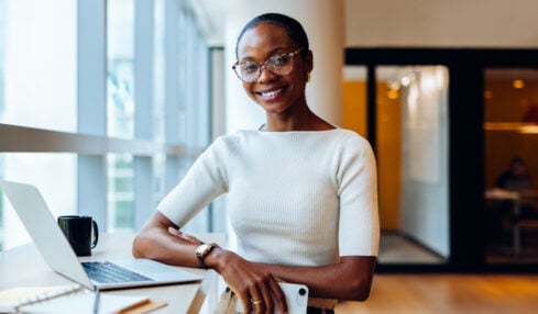 Young black businesswoman in an office environment, smiling confidently while using a laptop. She embodies a professional and approachable demeanor, reflective of productivity and success in business.