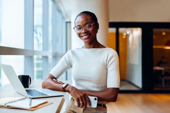 Young black businesswoman in an office environment, smiling confidently while using a laptop. She embodies a professional and approachable demeanor, reflective of productivity and success in business.